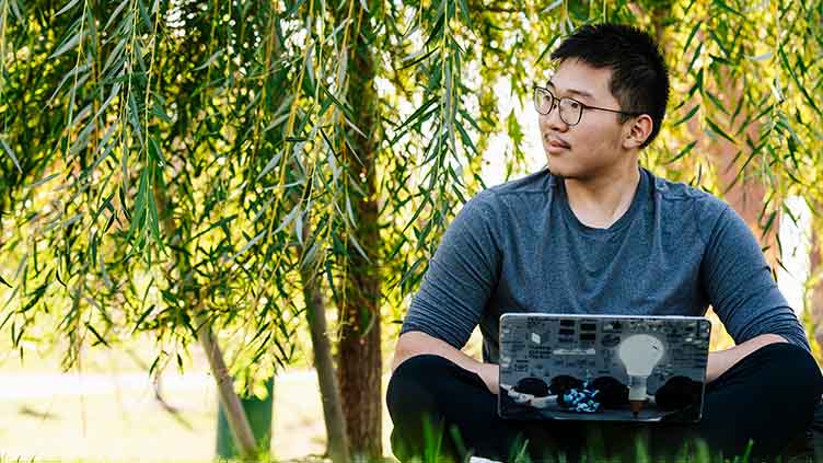 Summer 2025 timely update / male student sitting in the grass outside infront of the trees