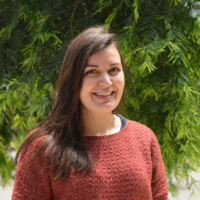 woman with long brown hair, reddish sweater, standing in front of greenery