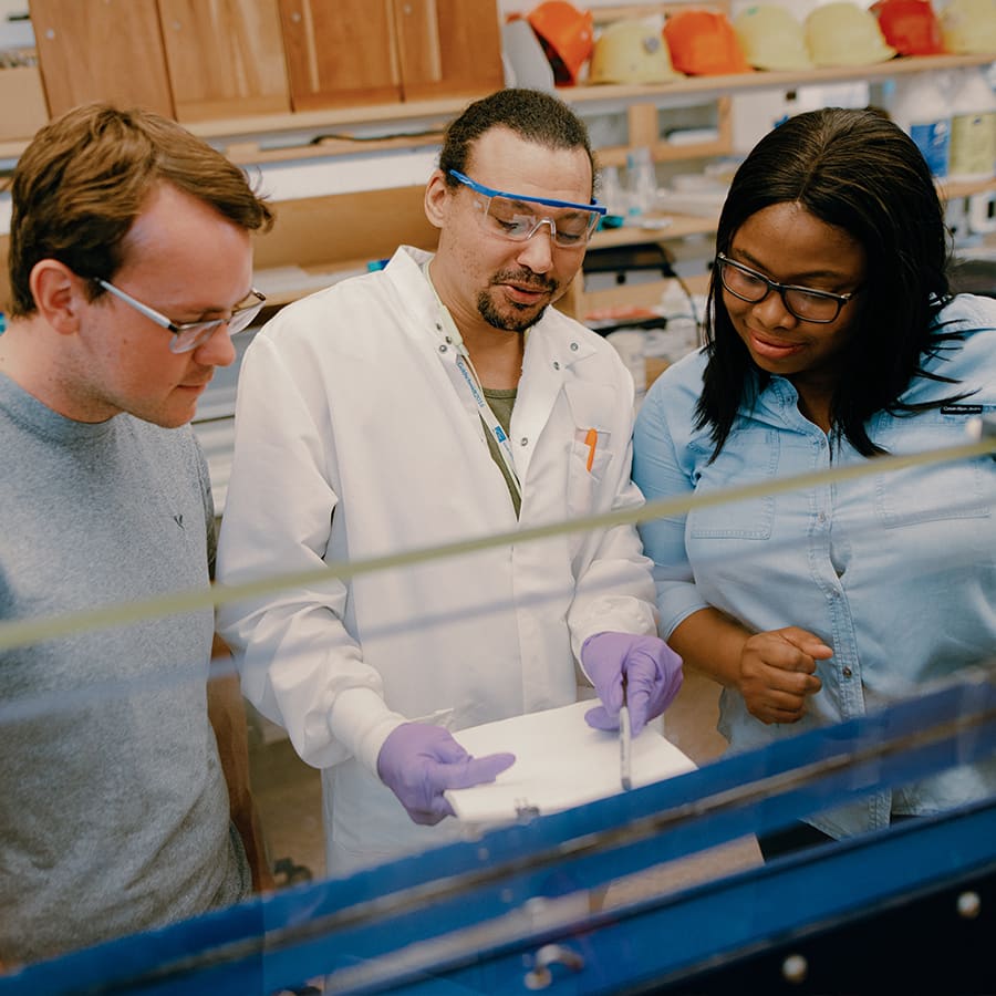 A grad student wearing a lab coat teaches 2 students in an environmental lab.